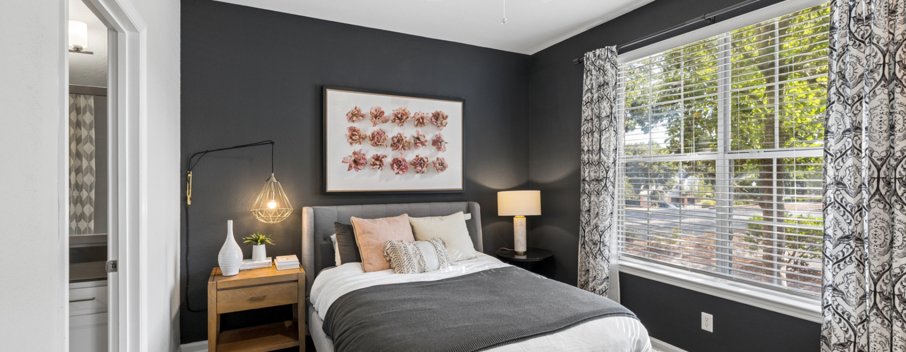 Modern apartment bedroom with dark accent walls, a large window, ceiling fan, upholstered bed and adjoining bathroom.